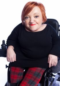 Woman with short red hair, sitting in wheelchair, smiling.